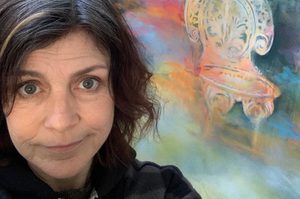 Angela is next to a colourful painting, with long black hair wearing a black coat