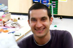 Benny is sat down, wearing a blue jumper and jeans with short dark hair