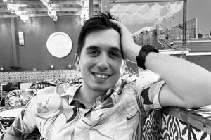 Ivo is leaning against a patterned wall. Ivo has short brown hair and is wearing a patterned shirt.