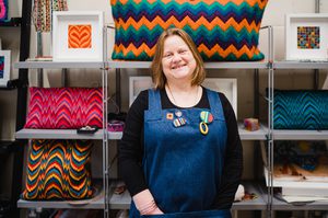 Tina is wearing blue dungarees and has a short brown bob. Tina is stood in front of colourful fabrics.