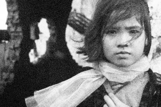 A Vietnamese girl stares directly into the camera, clutching a scarf around her neck. A bombed out building sits in the background.
