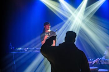 Man on stage with a microphone. There are white and blue strobe lights behind him. In front of the stage is a silouette of a man listening to the performer.