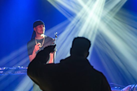 Man on stage with a microphone. There are white and blue strobe lights behind him. In front of the stage is a silouette of a man listening to the performer.