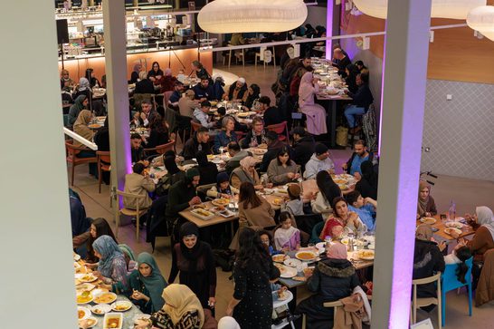 Image shows a softly lit restaurant filled with people sat at long rows of tables eating.