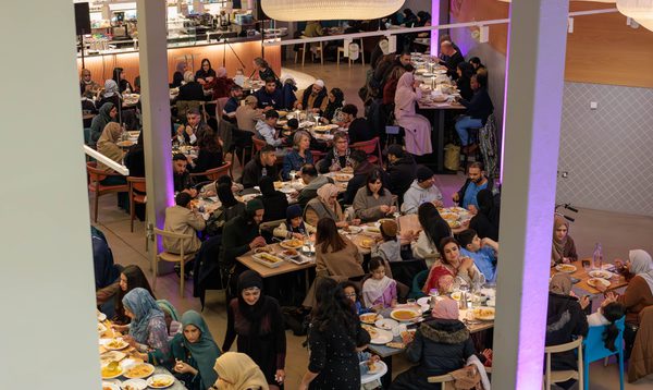 Image shows a softly lit restaurant filled with people sat at long rows of tables eating.