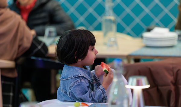 Image shows a small South Asian girl with a short bob cut and fringe in a denim dress. She is sitting in a restaurant space eating a slice of watermelon,
