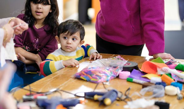 Image shows a group of people sitting at a table doing various textile crafts. In the centre of the image is a small South Asian boy in a green, white, yellow and red stripey jumper. Behind him is a small South Asian girl with mid-length black hair and a fringe, wearing a purple top.