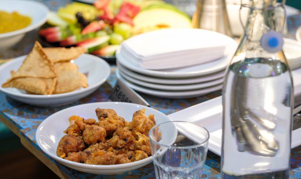 Image shows samosas and chicken pakoras on a blue patterned table, along with plates, napkins, cutlery, a bottle of water and fresh fruit.