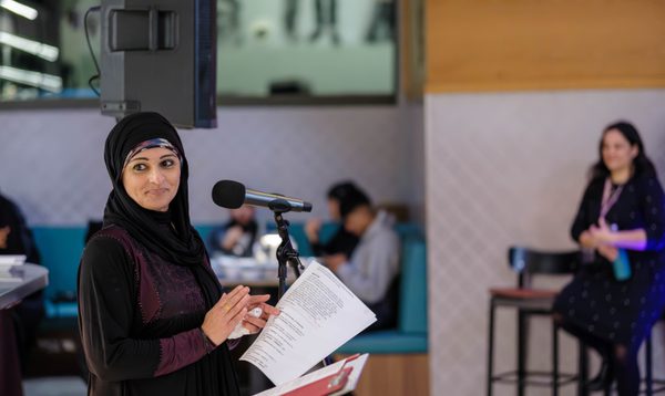 Image shows a South Asian woman in a black hijab and purple dress speaking at a microphone. She holds a piece of A4 paper with a speech written on it in her hand. In the background is a restaurant space where multiple people are seated.