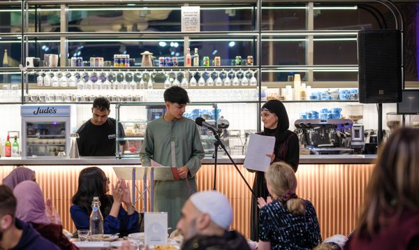 Image shows a softly lit restaurant space where two people (a young South Asian man and a South Asian woman) are making a speech at a microphone.