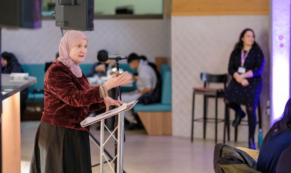 Image shows a white woman wearing a pale pink hijab, burgandy top and black skirt making a speech in front of a microphone and lecturn. Behind her is a restaurant space where people are seated.