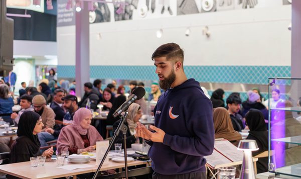 Image shows a young South Asian man with a beard and wearing a navy hoodie standing in front of a microphine. He is in the middle of singing the call to prayer, and hold his hands in front of him with palms facing up. Behind him is a crowded restaurant space.