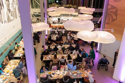 Image shows a softly lit restaurant filled with people sat at long rows of tables eating.