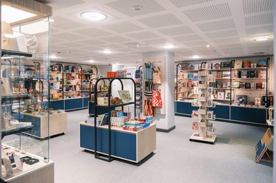 Image shows a gift shop filled with a variety of colourful art-related products. The floor of the shop is grey, while the pillars and ceiling are white. There is both glass and free standing light wood and blue display casing in the shop.