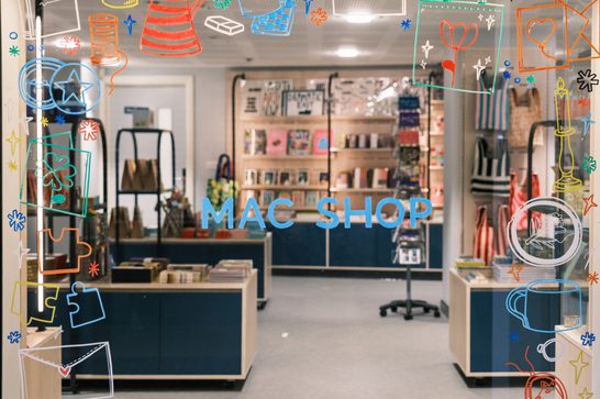 Image shows a glass shop window decorated with colourful window art. In the centre of the window are the words 'MAC Shop' in light blue capital letters, while the window art decorating the edges of the glass features various products sold in the shop. Behind the window is a gift shop filled with various art-related products.