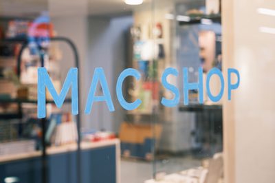 Image shows a shop window decorated with the words 'MAC Shop' in light blue capital letters. Behind the window art is a view of a gift shop stocked with various art-related products, glass display cabinets and dark blue and light word display stands.