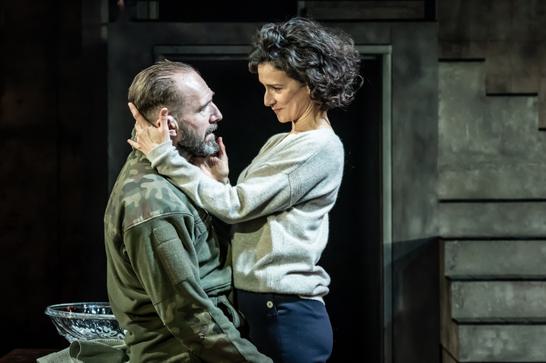 Dressed in camoflauge, Ralph Fiennes sits on the edge of a table whilst Indira Varma holds his head and strokes his face.