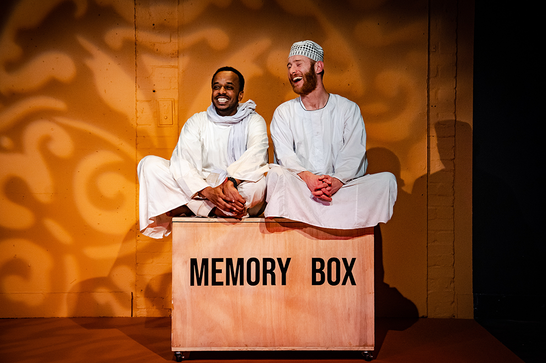 Mohand and Peter sit cross-legged and smiling on top of a box labelled "memory box".