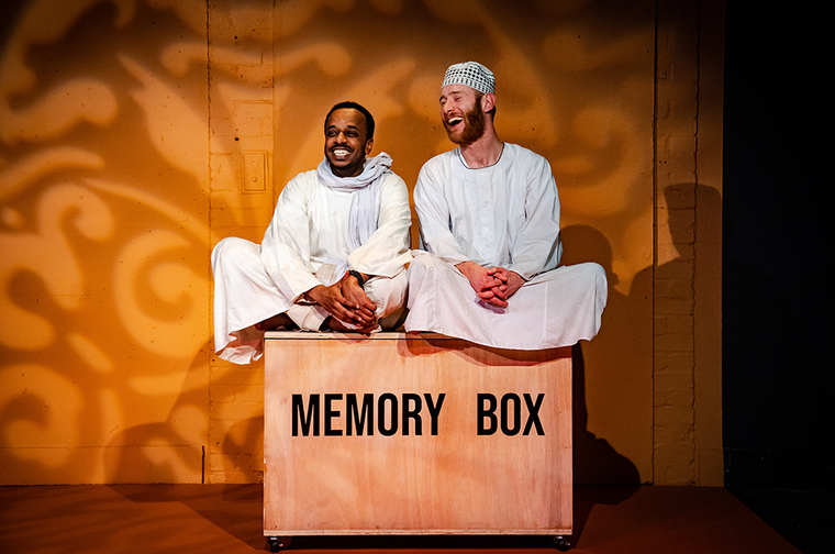 Mohand and Peter sit cross-legged and smiling on top of a box labelled "memory box".