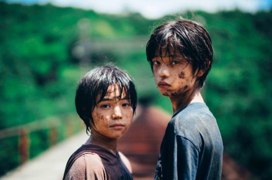 Two young Japanese boys covered in dirt look directly at the camera. A blurry background reveals that they are standing on train tracks.