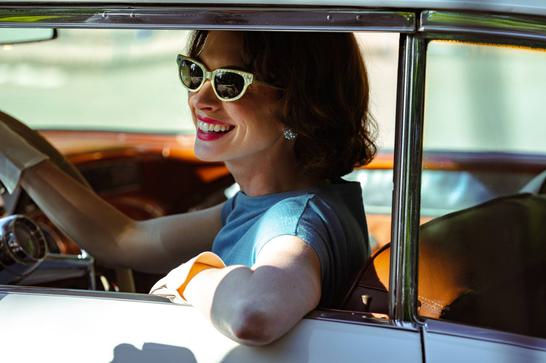 A smiling Anne Hathaway is sat in a 1950s car wearing sunglasses.