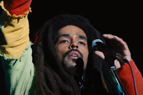 A man with dreadlocks (Bob Marley, as played by Kinglsey Ben-Adir) raises his hand, holding a microphone with the other hand.