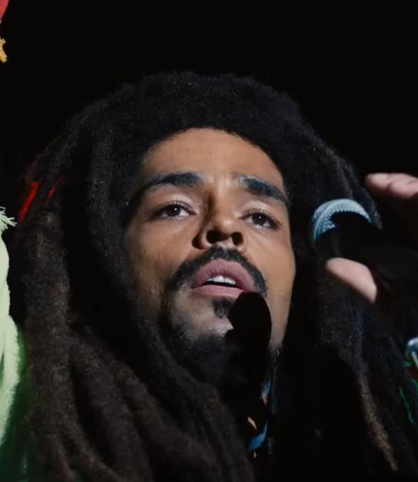 A man with dreadlocks (Bob Marley, as played by Kinglsey Ben-Adir) raises his hand, holding a microphone with the other hand.
