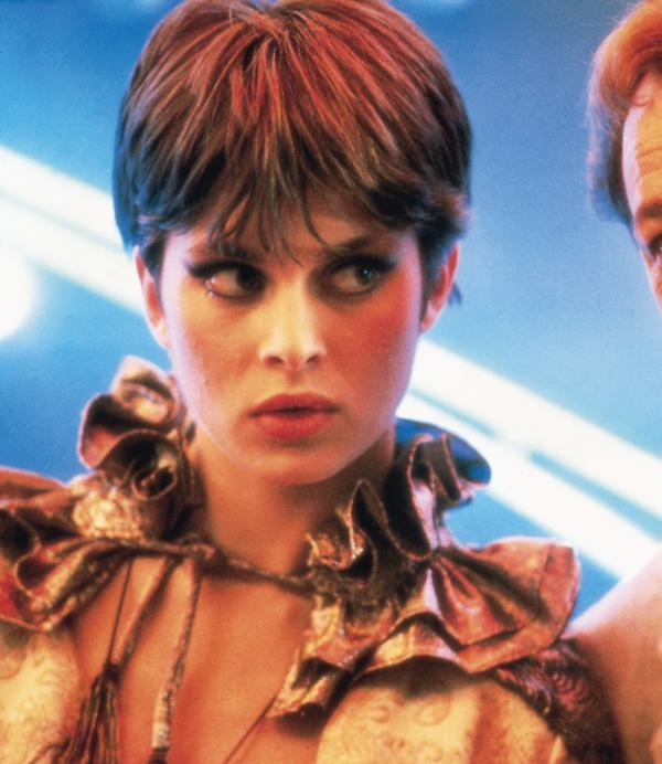 A man and a woman in 80s clothing stand side by side. The woman looks to her left, whilst the man looks directly into the camera.