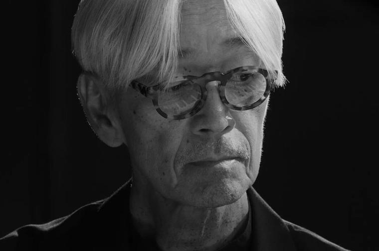 A simple black and white head shot of Ryuichi Sakamoto looking to his left.