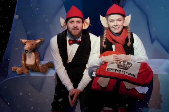 A fox and two of Santa's elves with a bag of letters sit on a snowy ledge and look forwards as snow falls.