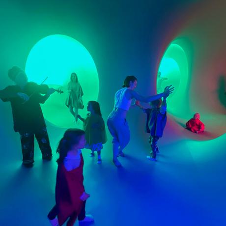 A violinist plays to several children and adults who move around a blue and green space with circular tunnels connecting to other chambers.
