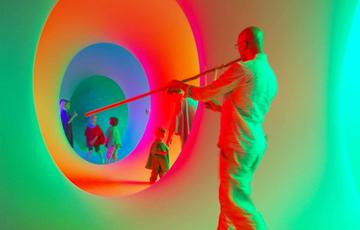 A man with his back to you plays a long pipe instrument and in the background you see children in a series of connected circular tunnels which are lit with bright colours.