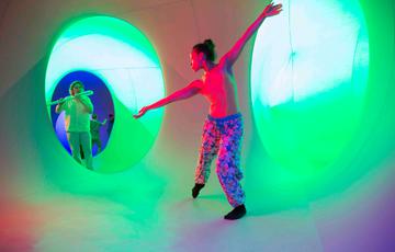 A woman dances with their arms spread out, and behind them another woman plays a flute-like instrument. The setting is a green and blue lit chamber with circular tunnels connecting to another chamber beyond.