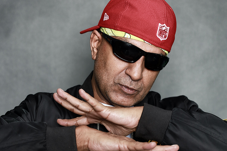 A man wearing a backwards red baseball cap and sunglasses (Apache Indian) crosses his arms with his fingers extended out.