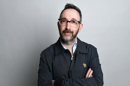 A man with glasses and a beard (Ashley Blaker) looks forward with his arms crossed in front of a plain grey background.