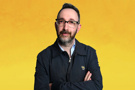 A man wearing glasses and with a beard (Ashley Blaker) looks forward with his arms crossed and a slight smile on his face in front of a yellow background.
