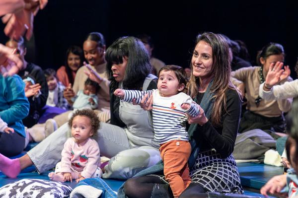 Parents and their babies smile watching a performance that is out of the shot. Babies look at the stage wide-eyed, sitting on their mum's knee, who is smiling.