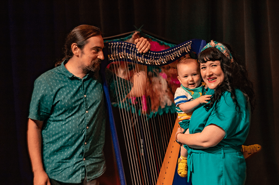 Two smiling adults, stood on a stage. One with an aquamarine jumpsuit and wavy black hair holds an infant. The other faces them, his arm over the harp in his turquoise button up shirt.