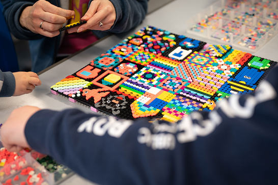 A close up of a patterned design made of Lego - you can see three people's hands and arms as they put more pieces of lego together.