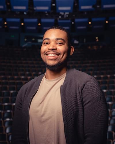 Man smiles at the camera in a beige top and grey cardigan. He is stood in a theatre auditorium.