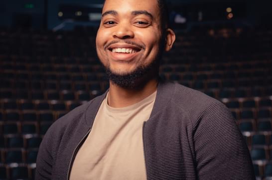 Man smiles at the camera in a beige top and grey cardigan. He is stood in a theatre auditorium.