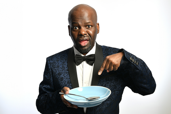 Daliso Chaponda wearing a tuxedo and pointing expectantly at an empty plate.