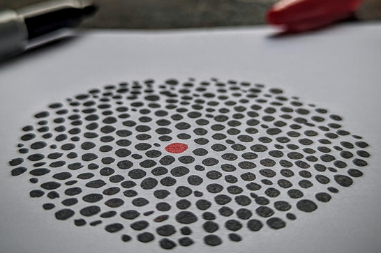 A red dot surrounded by black dots, drawn on paper with a black and red pens in the back pens in the background,