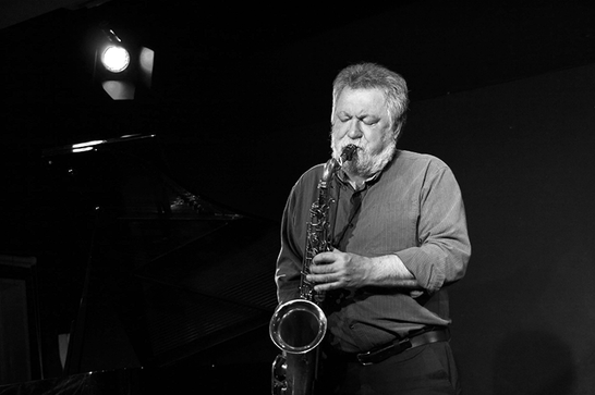 Black and white photo of a man with grey hair and a beard playing a saxophone.