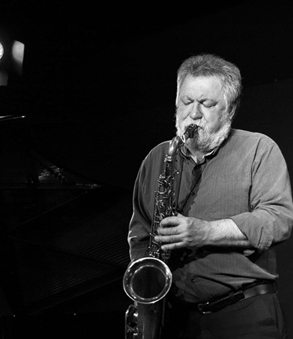 Black and white photo of a man with grey hair and a beard playing a saxophone.
