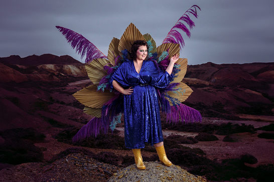A woman (Kiri Pritchard-McLean) stands outside in a sequined blue dress and with peacock feather around her.