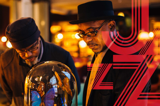 2 men in hats with glasses look at a butterfly in a glass domed case. The word JAZZ appears over the image.