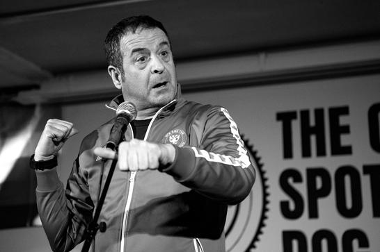Mark Thomas stands by a mic stand wearing a tracksuit top and gesturing with his fists.