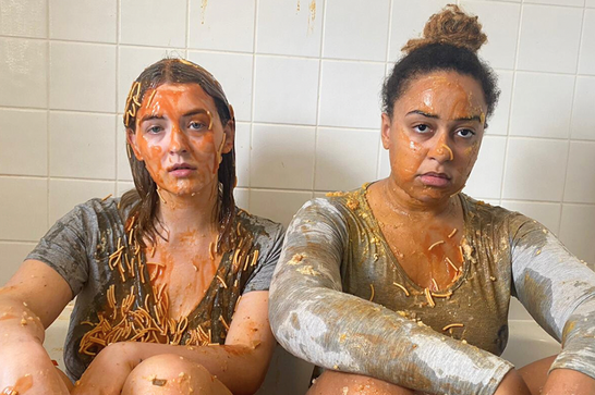 Two women sit in a bathroom with tinned spaghetti and tomato sauce all over their faces and clothes.