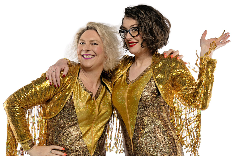 Two women - one with blond hair, another with dark hair and glasses - smile and wear shiny gold outfits with an arm round each other.